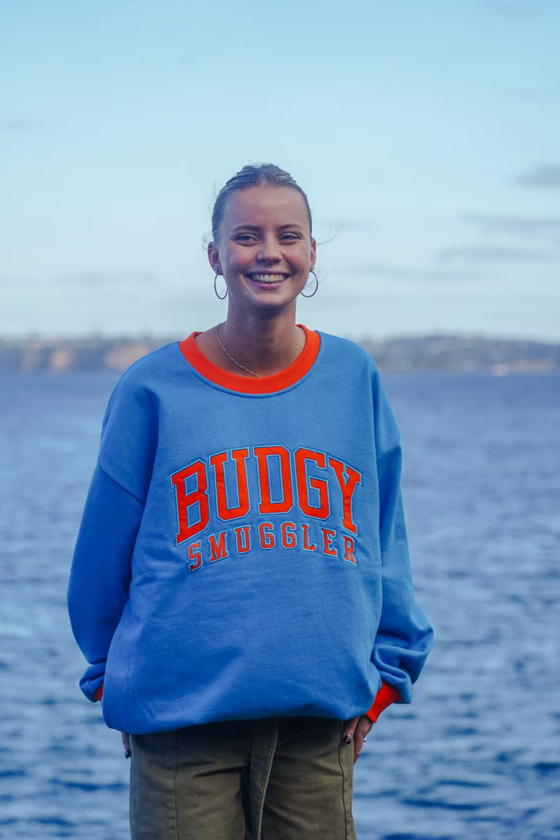 Person wearing a blue sweatshirt with 'BUDGY SMUGGLER' text standing by a body of water.