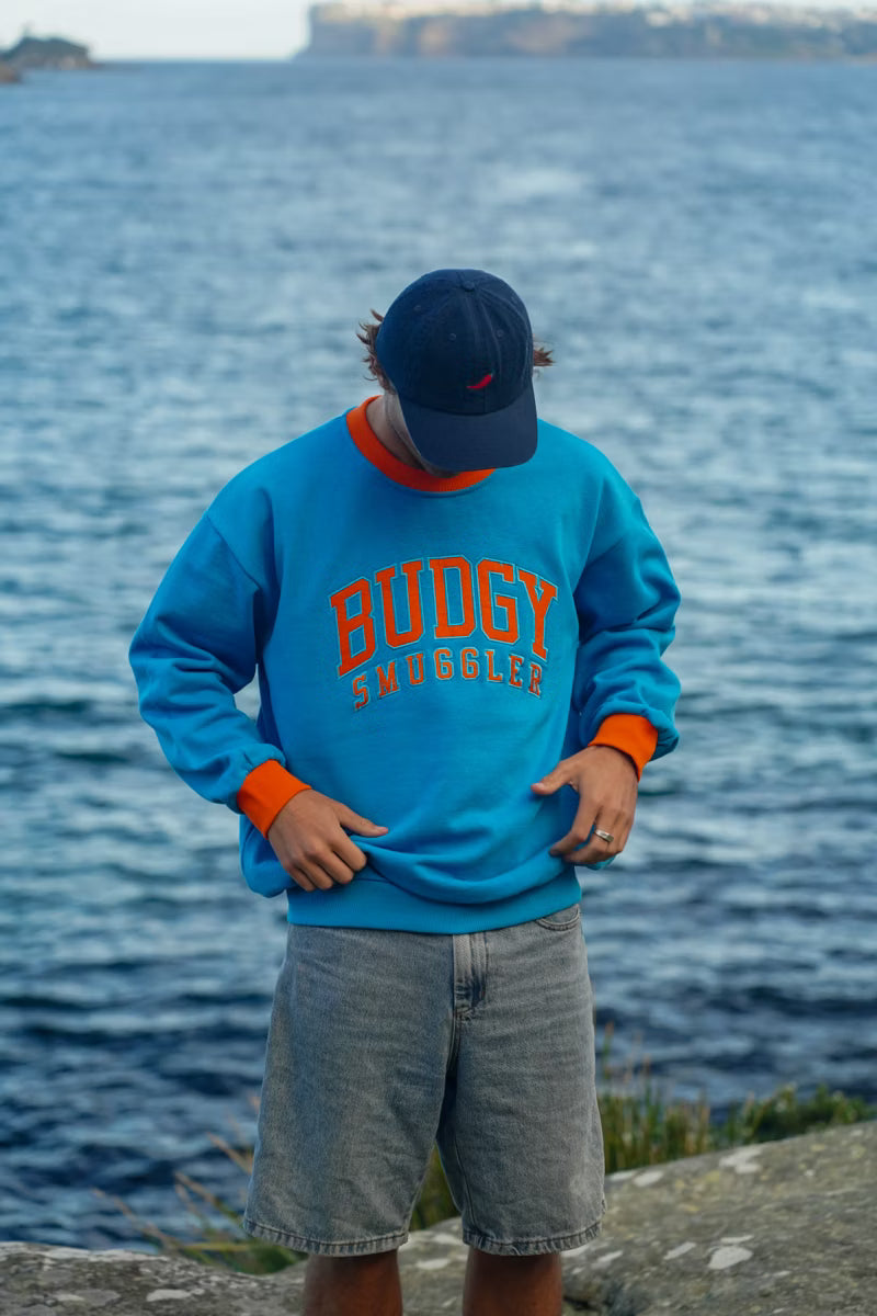 Person wearing a blue sweatshirt with 'Budgy Smuggler' text, standing by a body of water.