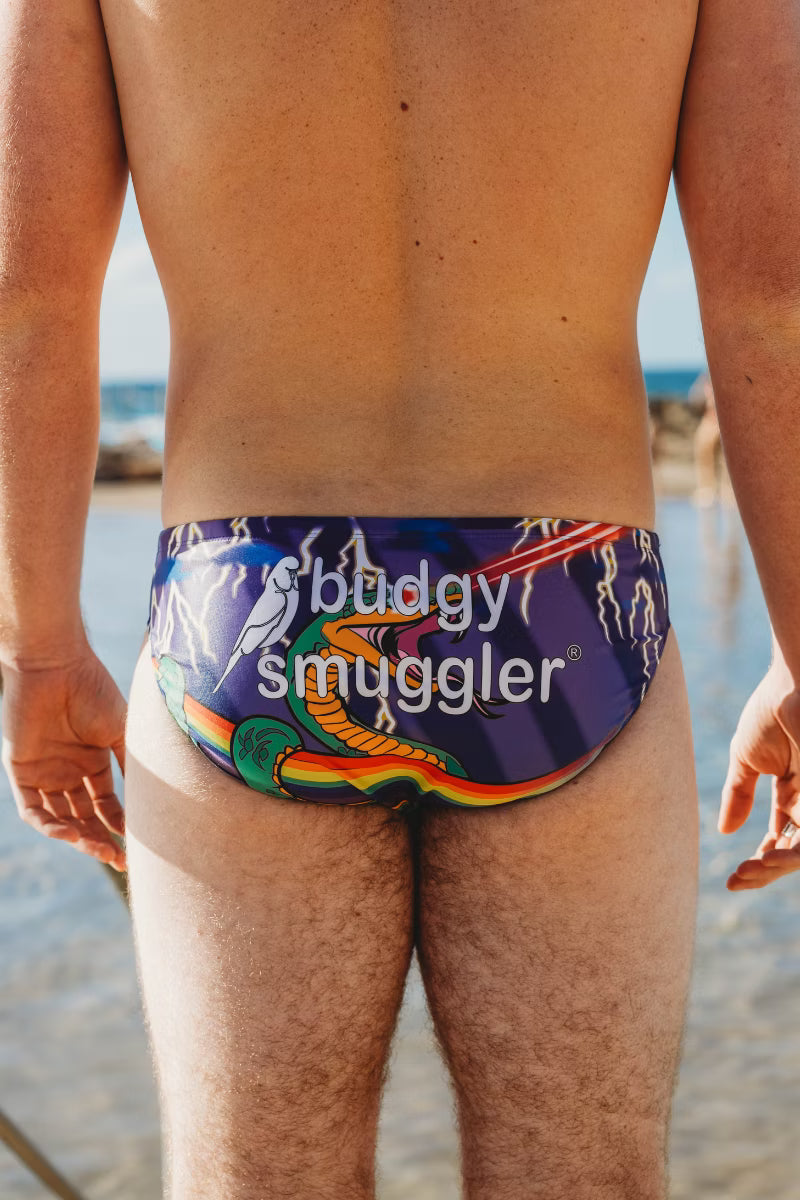 Person wearing a colorful swimsuit with 'budgy smuggler' branding on a beach.