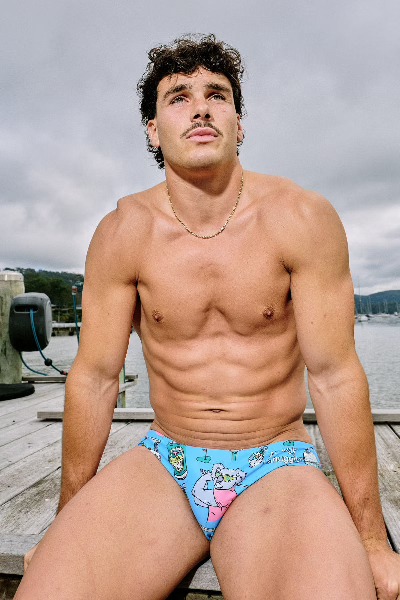 A shirtless man sitting on a wooden dock wearing light blue swim briefs with a cartoon bear pattern, with water and a cloudy sky in the background.